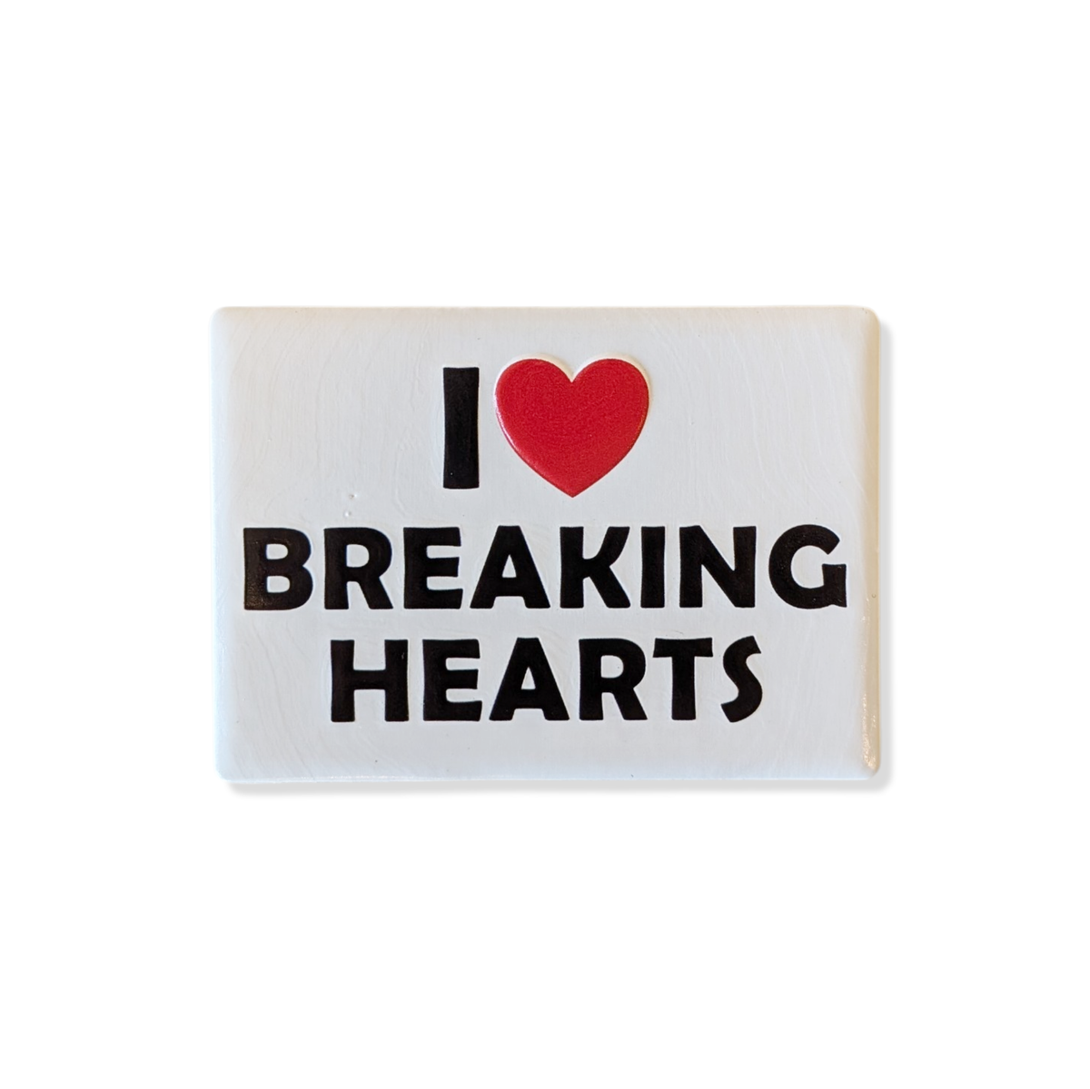 Rechteckiger Keramik-Kühlschrankmagnet (7x5 cm) im Querformat mit seidenmatter Glasur. Auf weißem Grund steht der schwarze Schriftzug 'I BREAKING HEARTS' mit einem leuchtend roten Herz-Symbol im fühlbaren 3D-Relief.