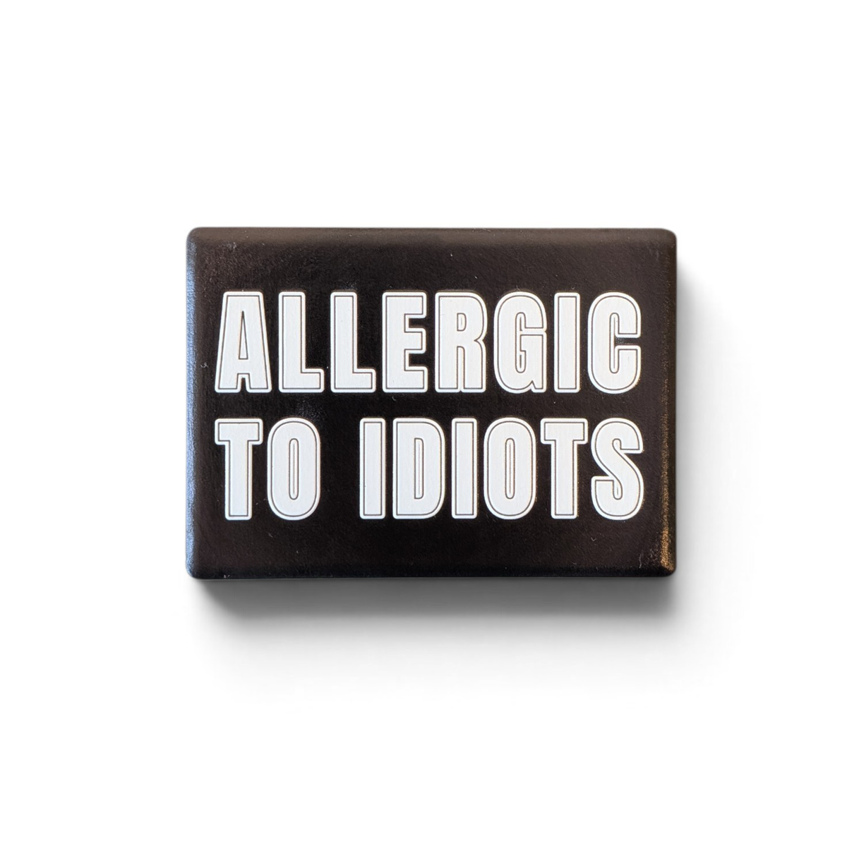 Rechteckiger Keramik-Kühlschrankmagnet (7x5 cm) im Querformat mit schwarzer, seidenmatter Glasur. Auf schwarzem Grund steht der weiße Schriftzug 'ALLERGIC TO IDIOTS' in doppelkonturierter Typografie im fühlbaren 3D-Relief.