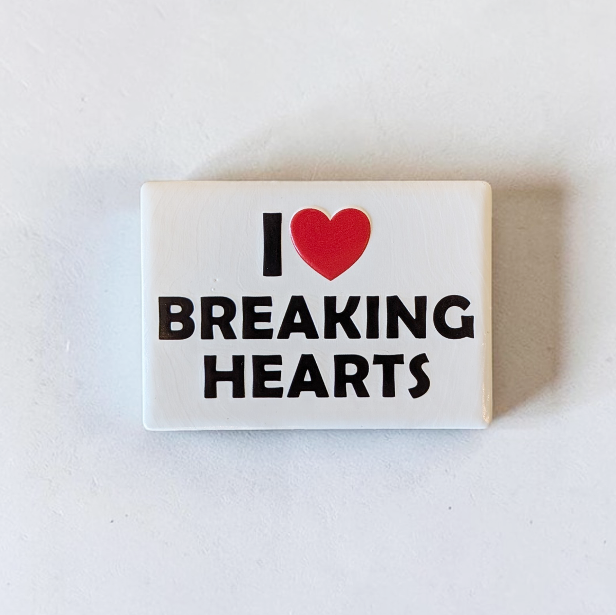 Rechteckiger Keramik-Kühlschrankmagnet (7x5 cm) im Querformat mit seidenmatter Glasur. Auf weißem Grund steht der schwarze Schriftzug 'I BREAKING HEARTS' mit einem leuchtend roten Herz-Symbol im fühlbaren 3D-Relief.