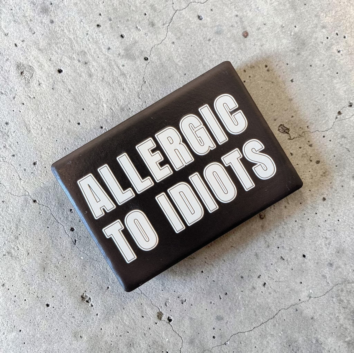 Rechteckiger Keramik-Kühlschrankmagnet (7x5 cm) im Querformat mit schwarzer, seidenmatter Glasur. Auf schwarzem Grund steht der weiße Schriftzug 'ALLERGIC TO IDIOTS' in doppelkonturierter Typografie im fühlbaren 3D-Relief.