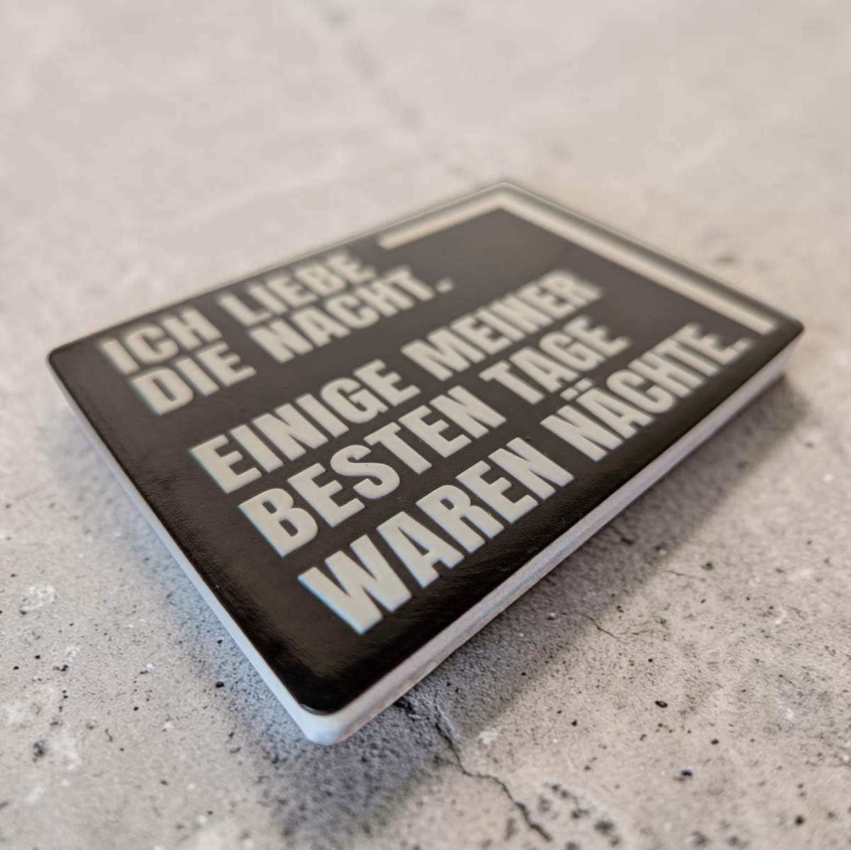 Rechteckiger Keramik-Kühlschrankmagnet (7x5 cm) im Querformat mit schwarzer, seidenmatter Glasur. Auf schwarzem Grund steht der weiße Schriftzug 'ICH LIEBE DIE NACHT. EINIGE MEINER BESTEN TAGE WAREN NÄCHSTE.' im fühlbaren 3D-Relief.