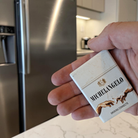 Rechteckiger Keramik-Kühlschrankmagnet (7x5 cm) mit seidenmatter Glasur im Design einer hellen Vintage-Zigarettenschachtel. Abgebildet sind Michelangelos Hände, die eine Zigarette weitergeben, mit dem Schriftzug 'MICHELANGELO SMOKING CONNECTS' im fühlbaren 3D-Relief.