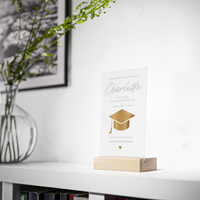 Transparenter Acrylglas-Aufsteller mit Holzsockel, personalisiert als Geschenk zum Abschluss. Zeigt goldene Doktorhut-Grafik, Namen, Studiengang und Hochschule in elegantem Design.
