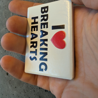 Rechteckiger Keramik-Kühlschrankmagnet (7x5 cm) im Querformat mit seidenmatter Glasur. Auf weißem Grund steht der schwarze Schriftzug 'I BREAKING HEARTS' mit einem leuchtend roten Herz-Symbol im fühlbaren 3D-Relief.