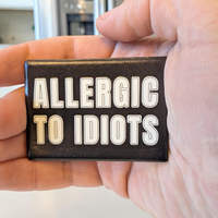 Rechteckiger Keramik-Kühlschrankmagnet (7x5 cm) im Querformat mit schwarzer, seidenmatter Glasur. Auf schwarzem Grund steht der weiße Schriftzug 'ALLERGIC TO IDIOTS' in doppelkonturierter Typografie im fühlbaren 3D-Relief.