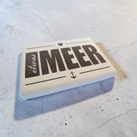Rechteckiger Keramik-Kühlschrankmagnet (7x5 cm) im Querformat mit seidenmatter Glasur. Auf weißem Grund steht der schwarze Schriftzug 'etwas MEER' mit Anker und Herz-Symbol im fühlbaren 3D-Relief.