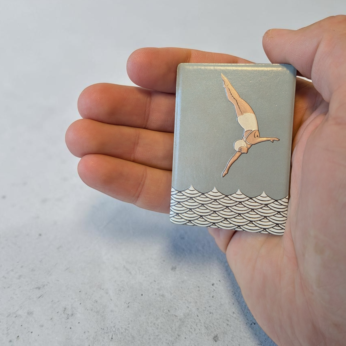 Rechteckiger Keramik-Kühlschrankmagnet (7x5 cm) mit seidenmatter Glasur in Salbeigrün. Zu sehen ist eine Turmspringerin im Art-Déco-Stil im Sprung über einem schwarz-weißen Wellenmuster im fühlbaren 3D-Relief.