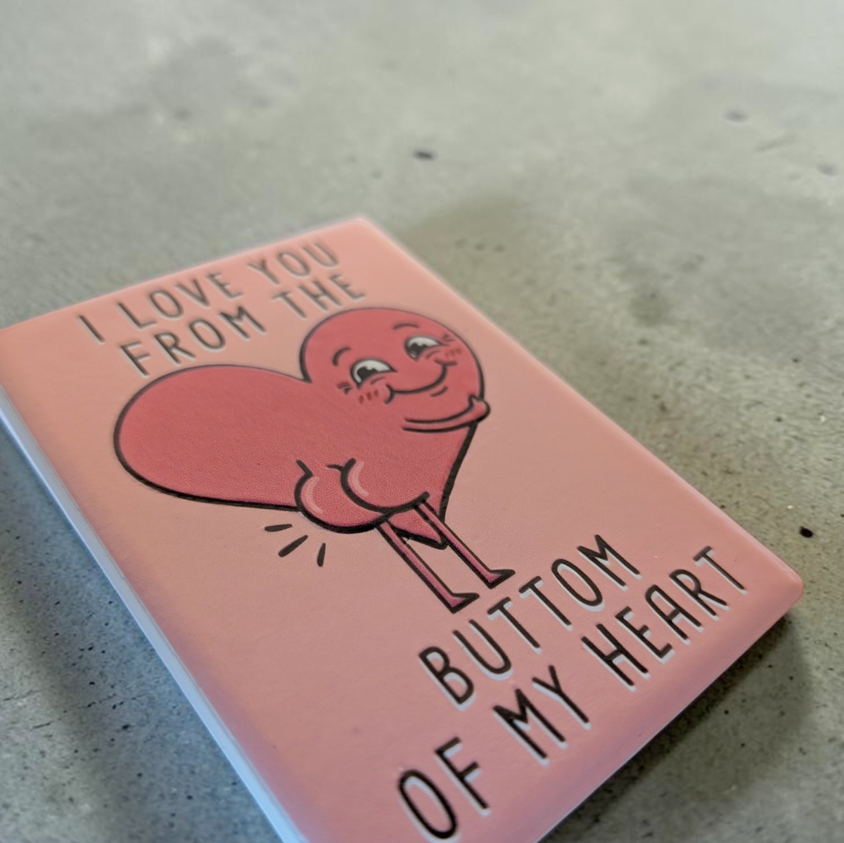 Rechteckiger Keramik-Kühlschrankmagnet (7x5 cm) mit seidenmatter Glasur in Altrosa. Abgebildet ist der schwarze Schriftzug 'I LOVE YOU FROM THE BUTTOM OF MY HEART' mit einem lustigen Herz inklusive Po im fühlbaren 3D-Relief.