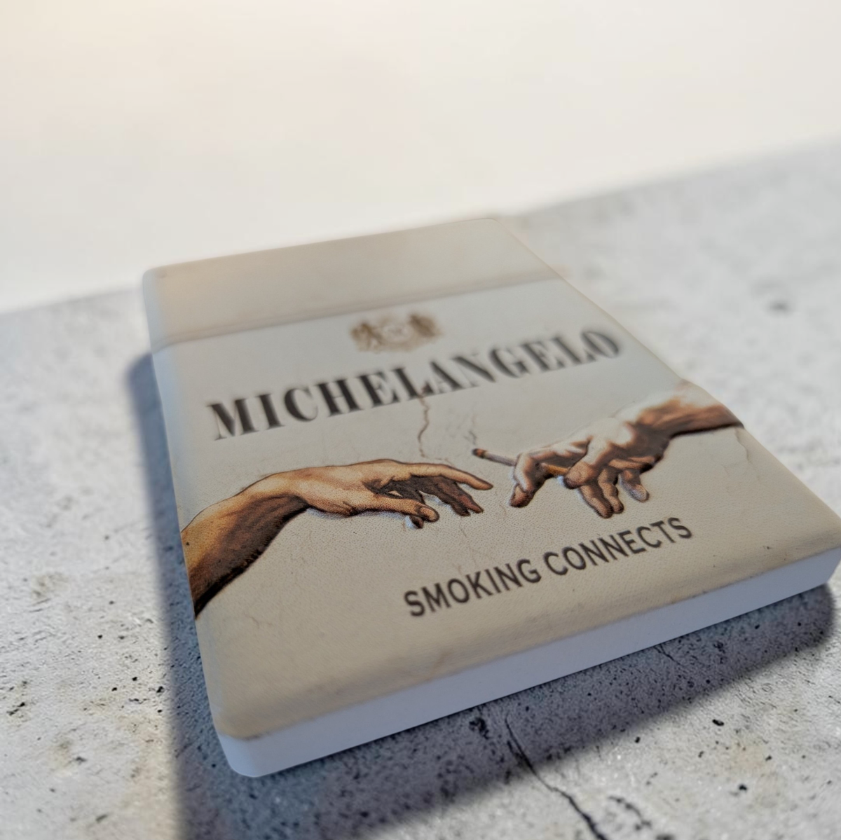 Rechteckiger Keramik-Kühlschrankmagnet (7x5 cm) mit seidenmatter Glasur im Design einer hellen Vintage-Zigarettenschachtel. Abgebildet sind Michelangelos Hände, die eine Zigarette weitergeben, mit dem Schriftzug 'MICHELANGELO SMOKING CONNECTS' im fühlbaren 3D-Relief.