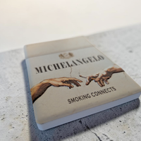 Rechteckiger Keramik-Kühlschrankmagnet (7x5 cm) mit seidenmatter Glasur im Design einer hellen Vintage-Zigarettenschachtel. Abgebildet sind Michelangelos Hände, die eine Zigarette weitergeben, mit dem Schriftzug 'MICHELANGELO SMOKING CONNECTS' im fühlbaren 3D-Relief.