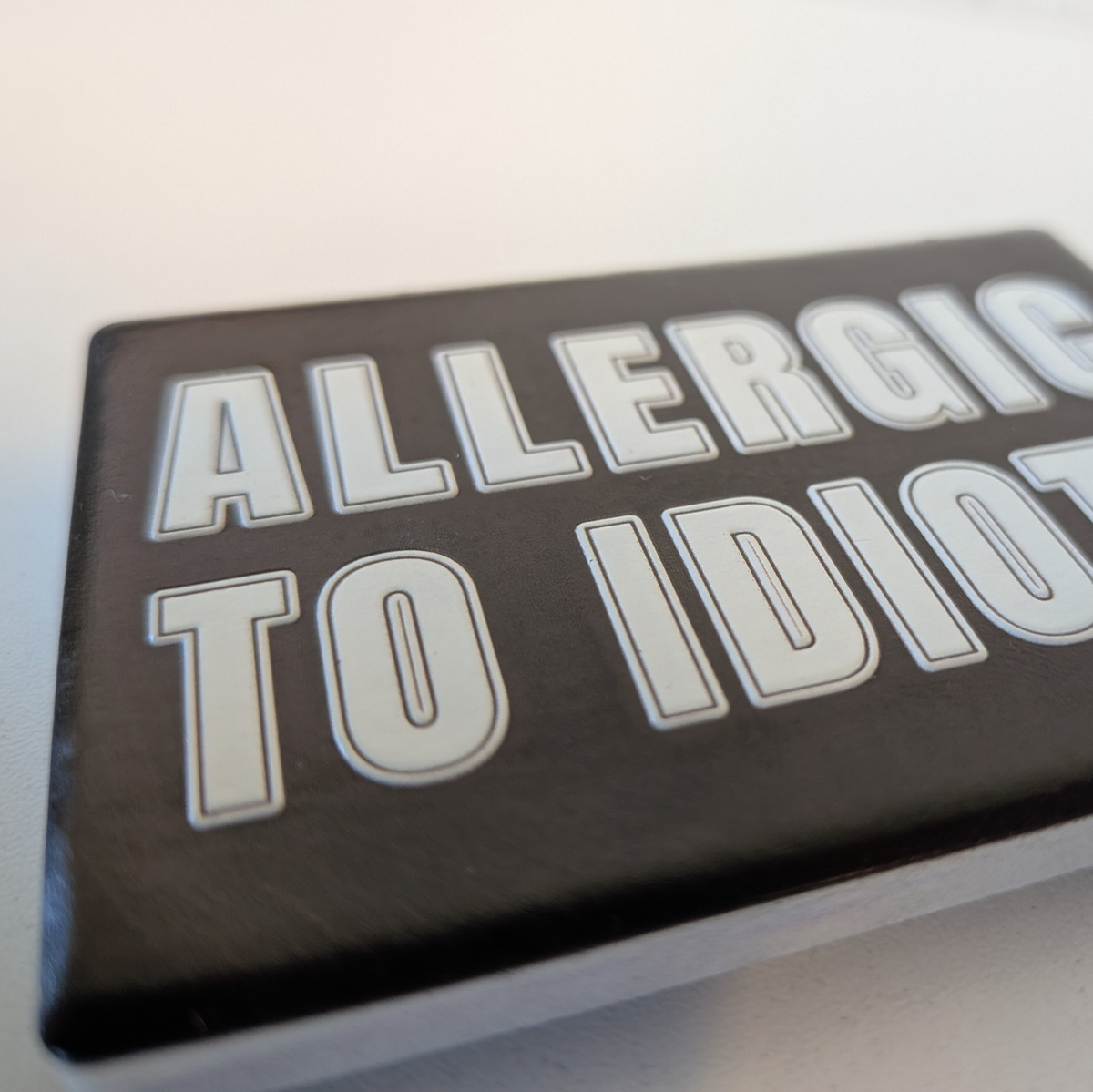 Rechteckiger Keramik-Kühlschrankmagnet (7x5 cm) im Querformat mit schwarzer, seidenmatter Glasur. Auf schwarzem Grund steht der weiße Schriftzug 'ALLERGIC TO IDIOTS' in doppelkonturierter Typografie im fühlbaren 3D-Relief.