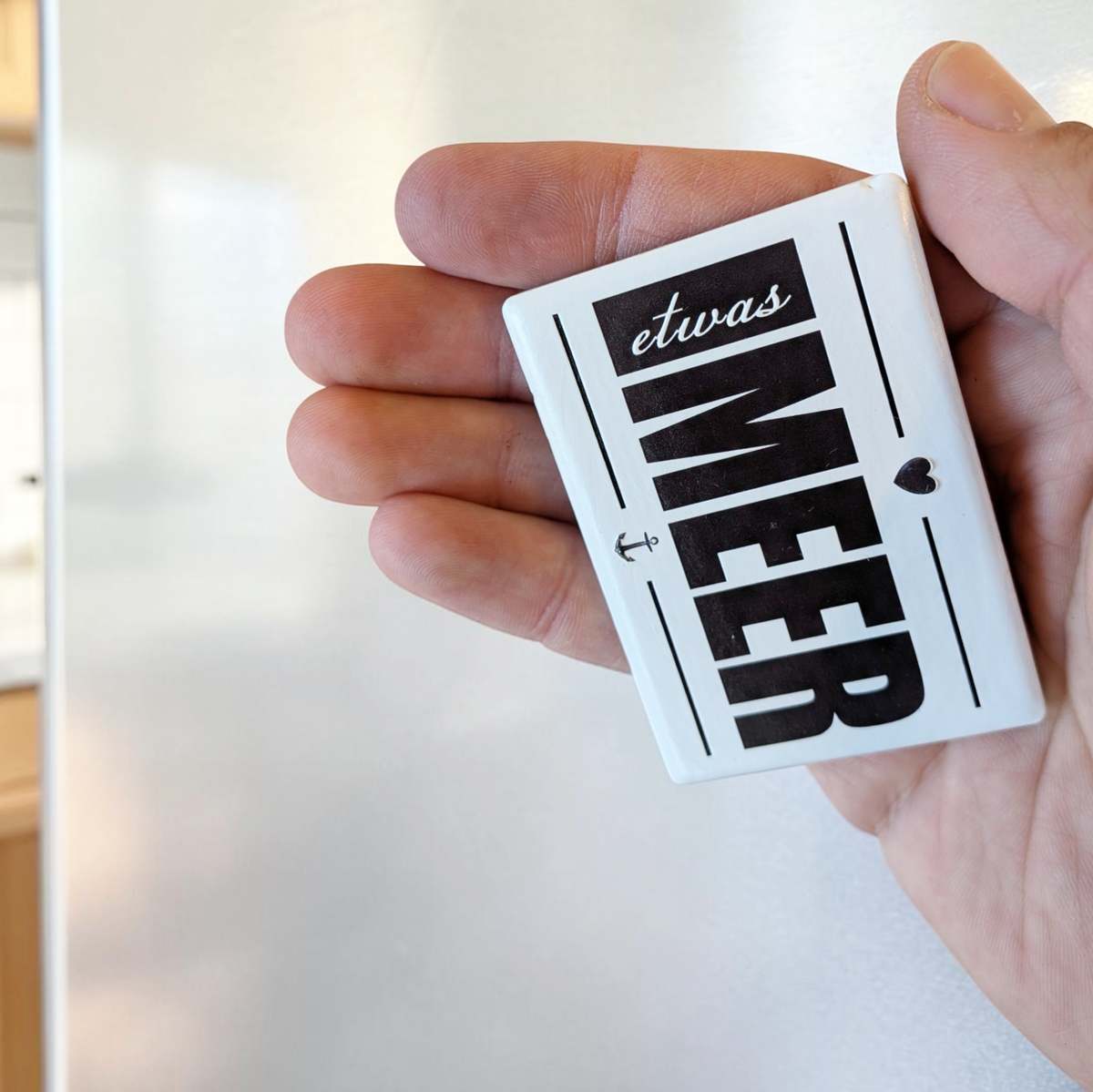 Rechteckiger Keramik-Kühlschrankmagnet (7x5 cm) im Querformat mit seidenmatter Glasur. Auf weißem Grund steht der schwarze Schriftzug 'etwas MEER' mit Anker und Herz-Symbol im fühlbaren 3D-Relief.