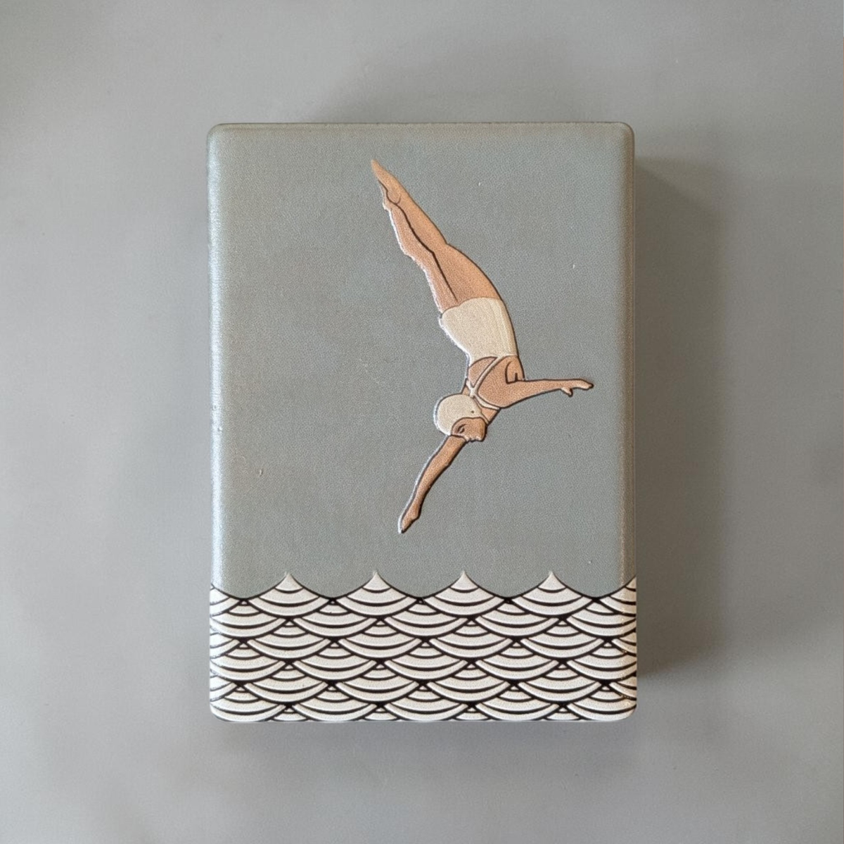 Rechteckiger Keramik-Kühlschrankmagnet (7x5 cm) mit seidenmatter Glasur in Salbeigrün. Zu sehen ist eine Turmspringerin im Art-Déco-Stil im Sprung über einem schwarz-weißen Wellenmuster im fühlbaren 3D-Relief.
