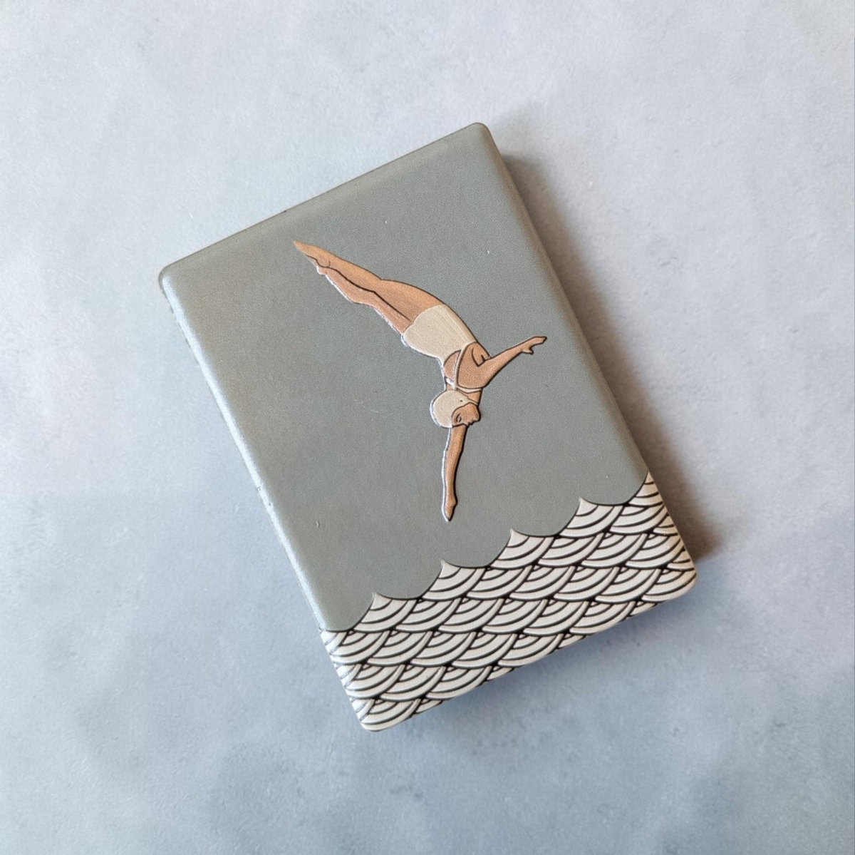Rechteckiger Keramik-Kühlschrankmagnet (7x5 cm) mit seidenmatter Glasur in Salbeigrün. Zu sehen ist eine Turmspringerin im Art-Déco-Stil im Sprung über einem schwarz-weißen Wellenmuster im fühlbaren 3D-Relief.